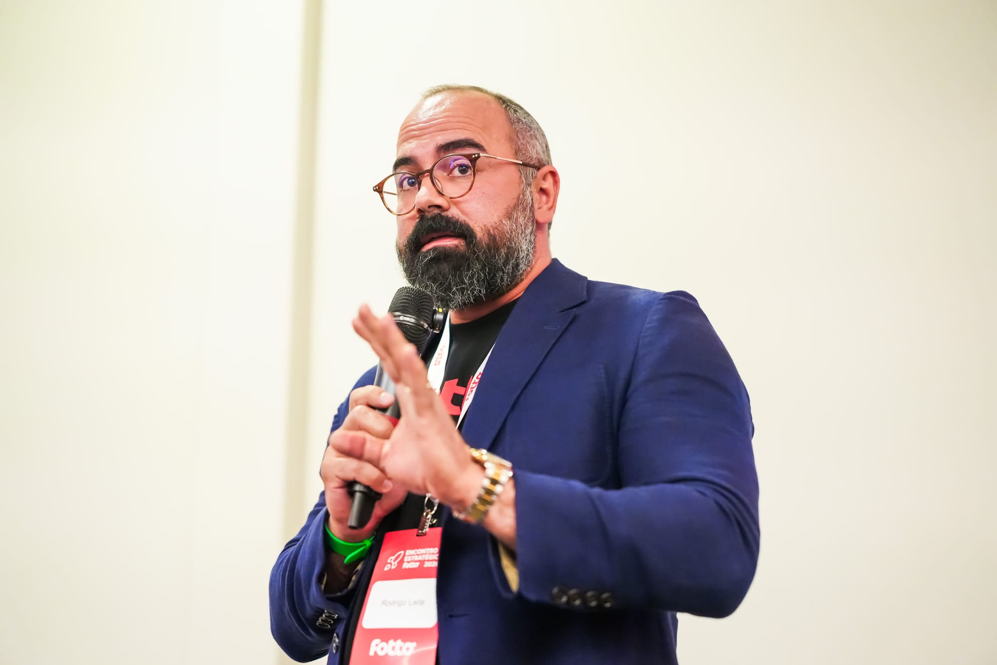 Homem de óculos fala ao microfone em uma apresentação. Ele veste blazer azul e crachá com o nome Rodrigo Leite e o logo da Fotto. O fundo é claro e neutro.