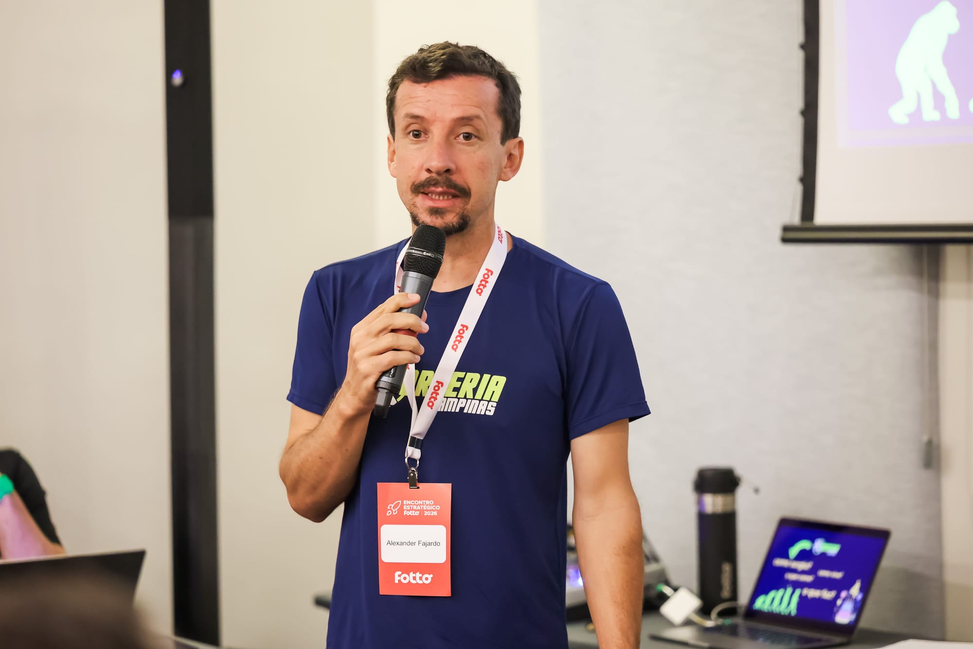 Homem com camiseta azul segura um microfone e fala durante uma apresentação. Ele usa crachá com o nome "Alexander Fajardo" e ao fundo há um notebook e um telão com projeção.