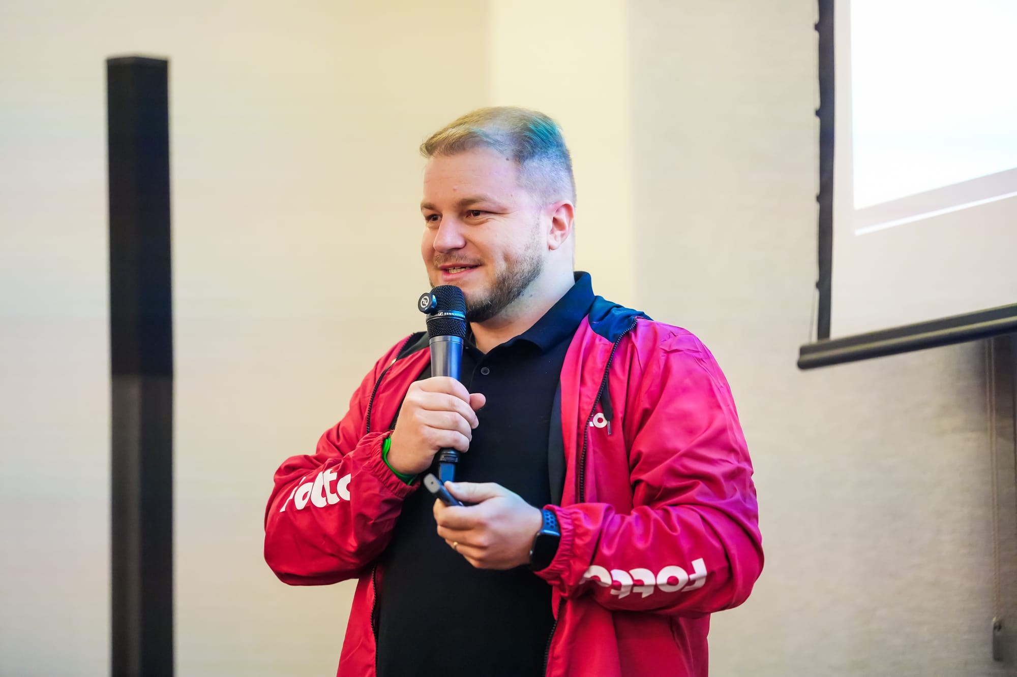 Homem loiro fala ao microfone durante apresentação. Ele veste uma jaqueta vermelha da Fotto e camisa preta. Ao fundo, tela de projeção e parede clara.