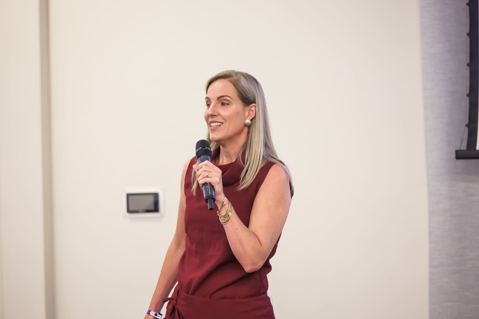 Mulher segurando microfone durante palestra em ambiente fechado, usando vestido vinho e crachá do evento Fotto.