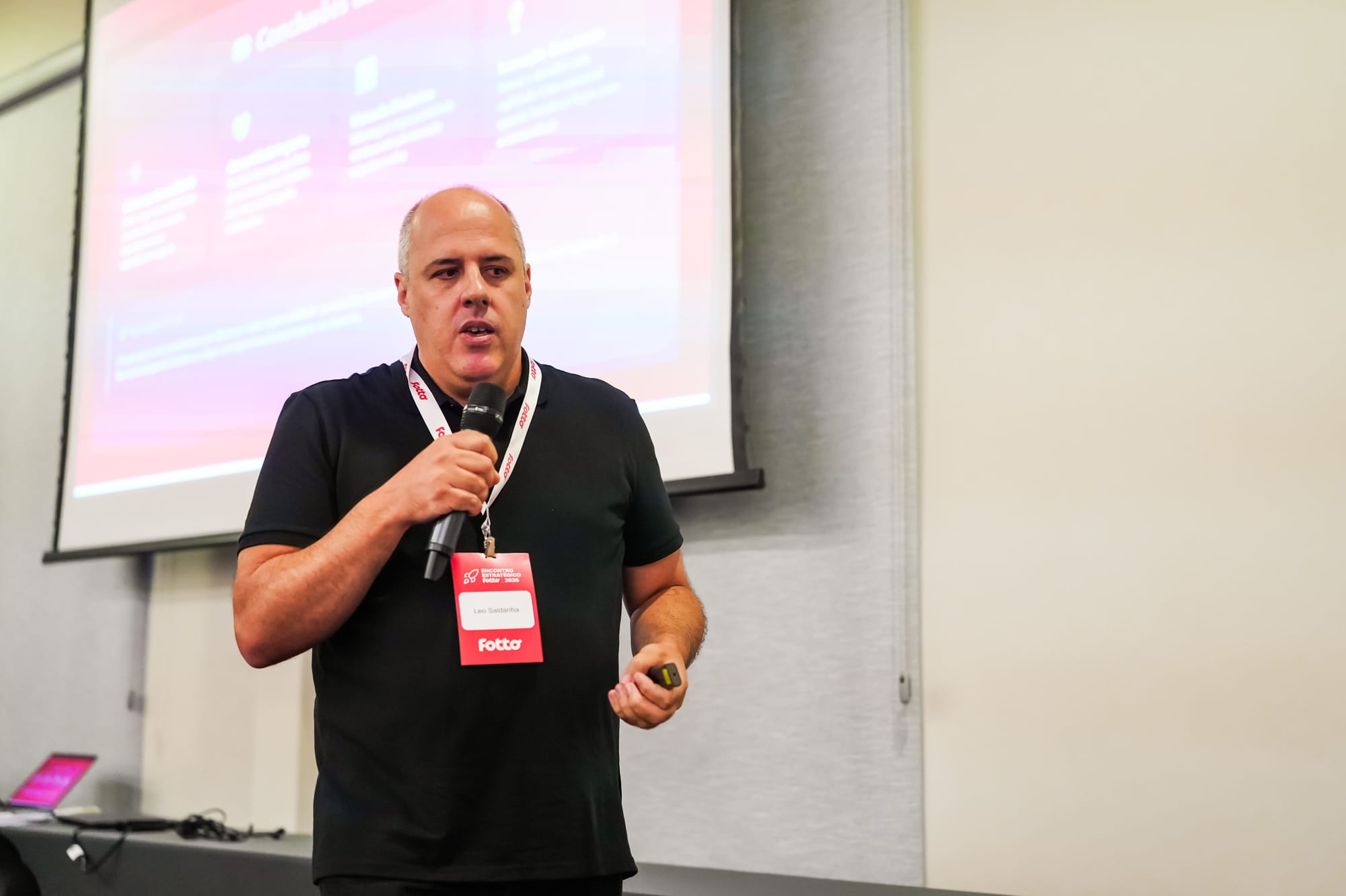 Homem segura um microfone e apresenta uma palestra em um ambiente fechado. Ele usa crachá com o nome "Leo Saldanha" e o logo da empresa "Fotto", com um telão de slides projetado ao fundo.