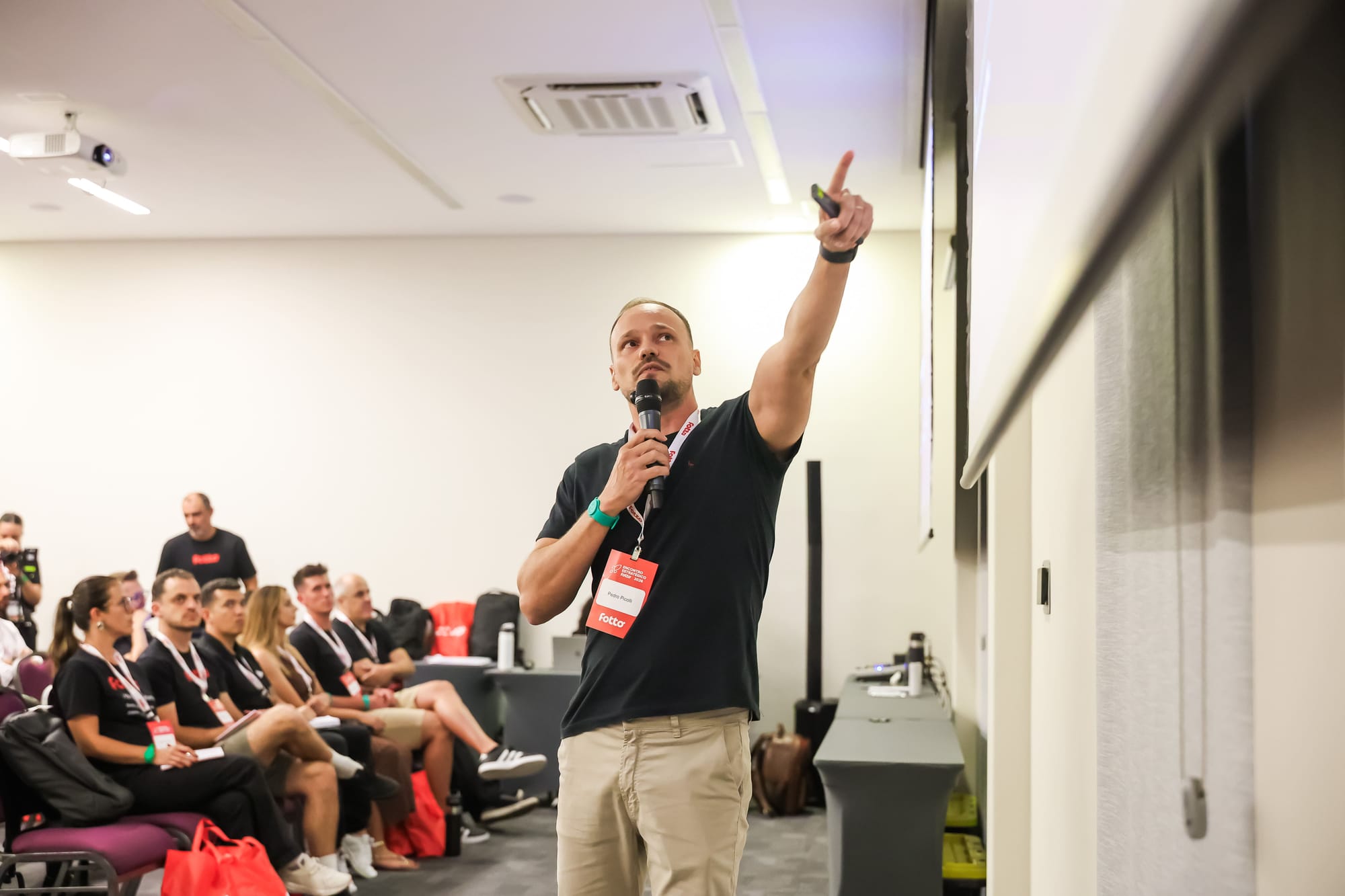 Homem de camiseta preta fala ao microfone e aponta para a tela durante apresentação. Ele está em pé em frente a um grupo de pessoas sentadas que assistem à palestra.
