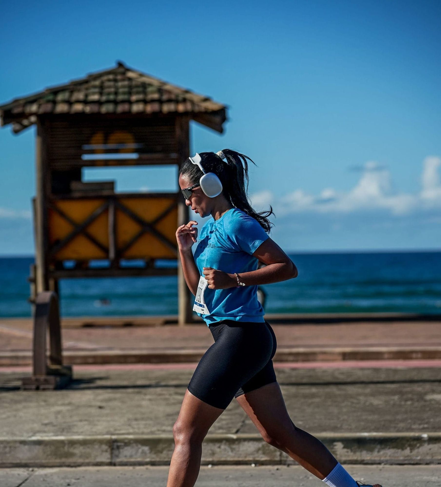 Mulher correndo na orla com fone de ouvido, usando roupa esportiva e número da prova preso à camisa.