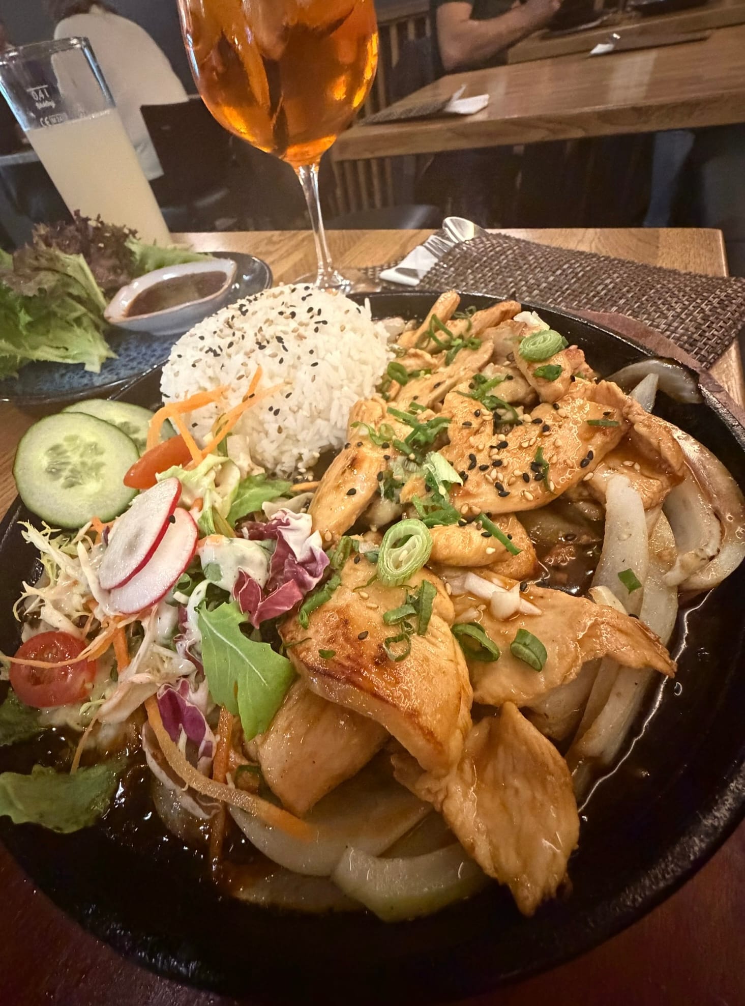 Dak Galbi auf heißer gusseiserner Platte: gebratene Hähnchenstreifen mit Zwiebeln in dunkler Sauce, garniert mit Frühlingszwiebeln und Sesam, dazu geformter Reis mit schwarzem Sesam und gemischter Blattsalat mit Gurke, Radieschen und Tomaten; im Hintergrund ein Glas Aperol Spritz © Nicole Isensee