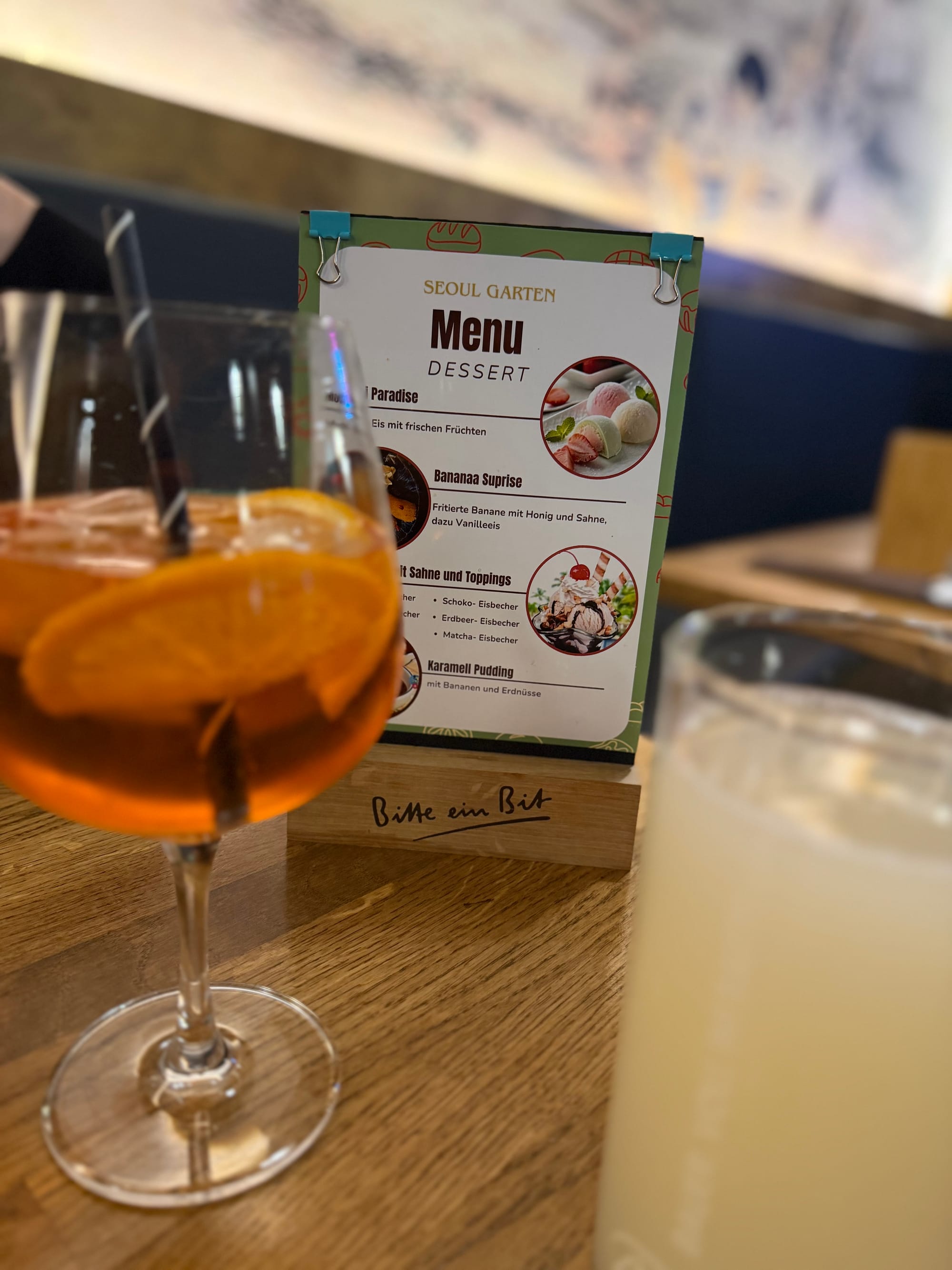 Dessert-Menükarte des Seoul Garten auf dem Tisch, davor ein Aperol Spritz und mit Orangenscheibe und einem Glas Litschisaft © Roman Jasiek