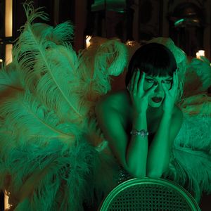 Alt-Text: Person im Glamour-Outfit mit Federboa lehnt nach vorne, Gesicht verdeckt, grünliches Licht. Urheberrechte liegen bei Universal Music.