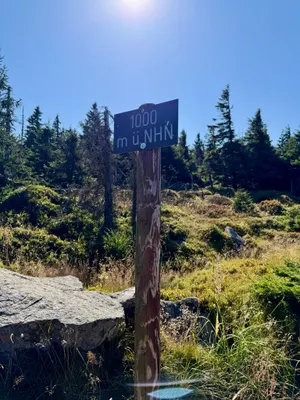 Ein hölzerner Wegweiser mit einem dunkelblauen Schild und der Aufschrift 1000 m ü. NHN vor einer weiten Berglandschaft im Harz. © Roman Jasiek