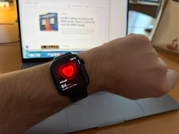 Apple Watch am Handgelenk zeigt die Herzfrequenz-App mit 84 BPM, im Hintergrund ein MacBook mit geöffnetem Blog (© Roman Jasiek)