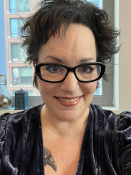 A photo of the author, a smiling woman with spiky black hair and glasses. She is wearing a grey-blue velvet top, and the face and beak of a tattooed raven is peaking out of it.