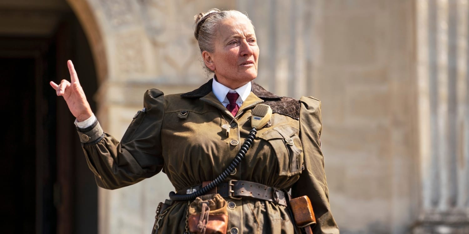 A massive woman with a tight grey bun wearing military-inspired garb gestures dramatically.