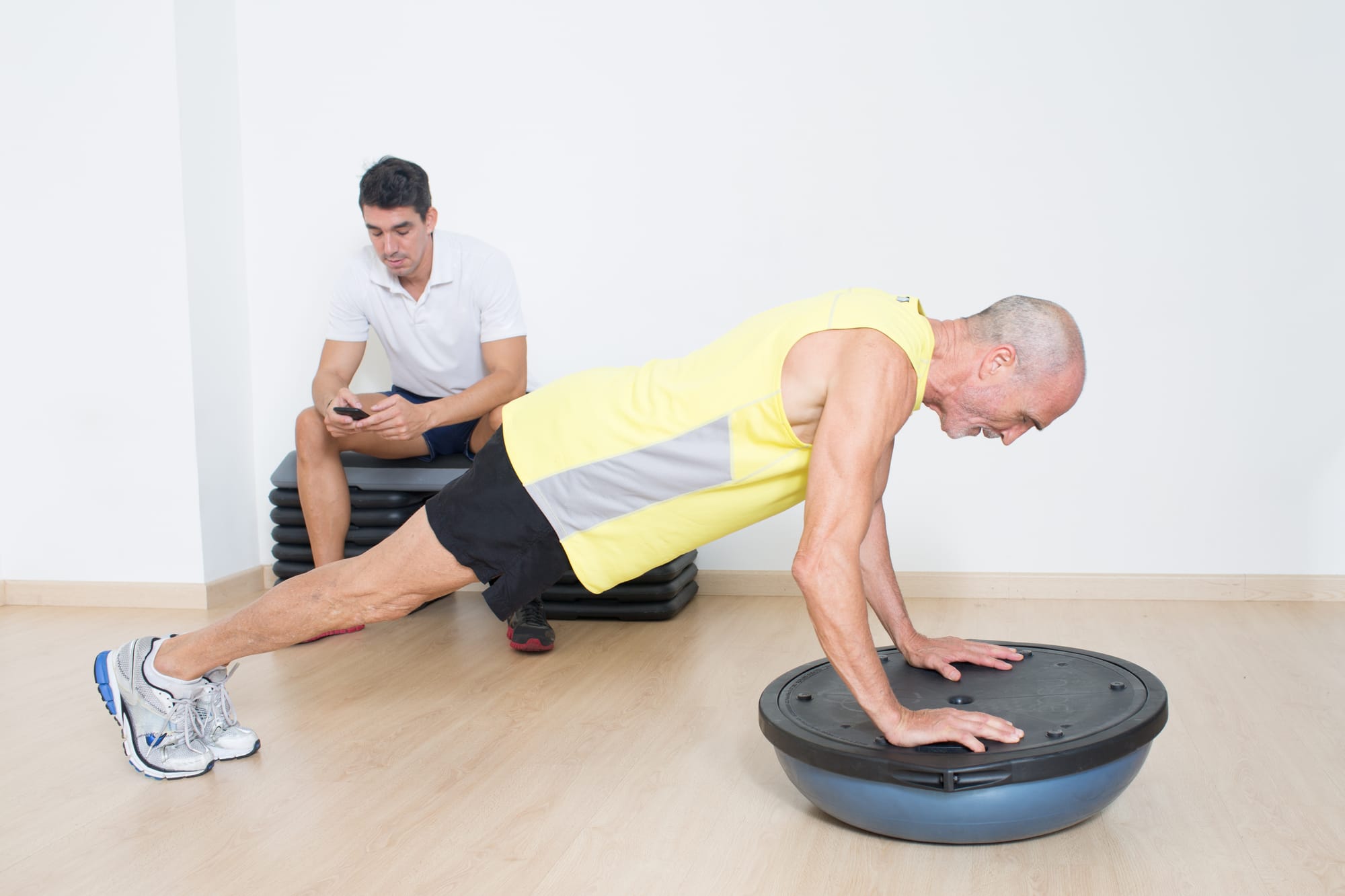 A fitness client performs a risky balance move while his trainer checks his email on his phone