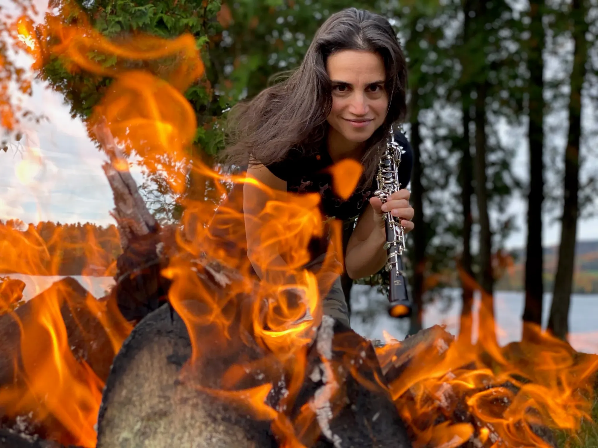 A photo of oboist Katherine Needleman. A fierce, smiling woman with long, deep brown hair brandishes an oboe outdoors with billowing flaming in the foreground, a lake and trees in the background.
