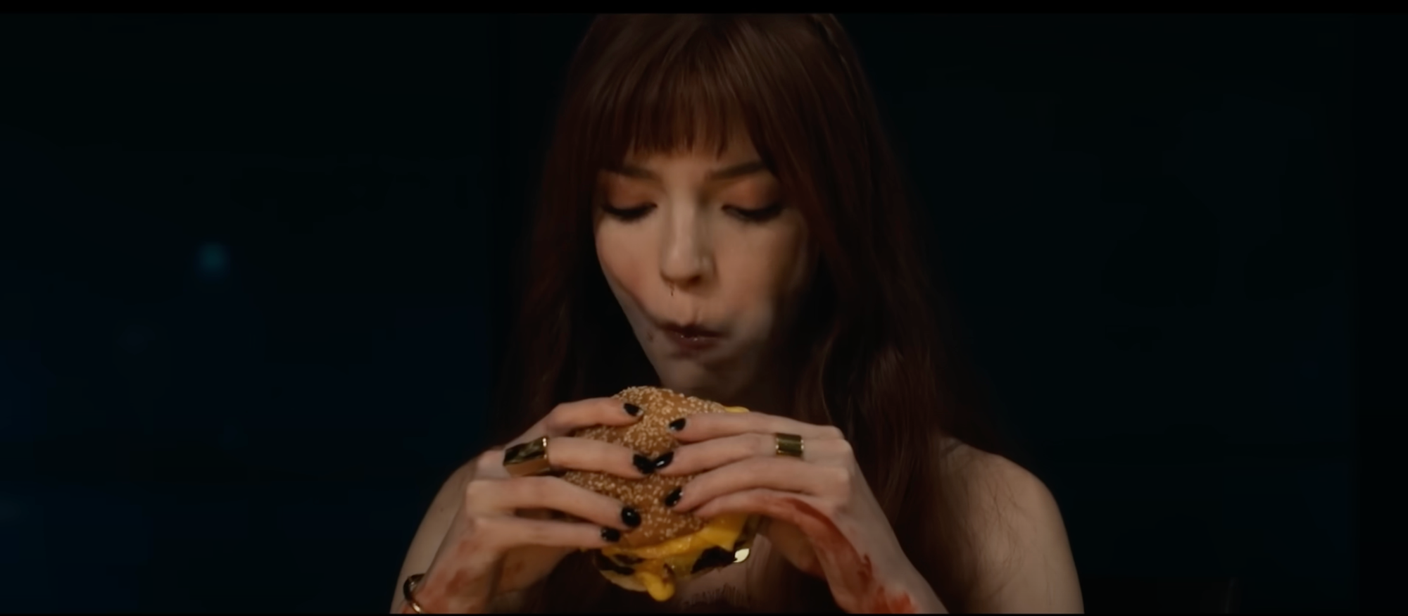 A still from The Menu. A young woman played by Anya Taylor-Joy, takes a bite of the best cheeseburger ever served.