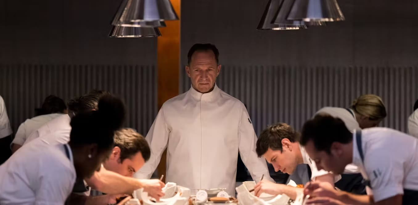 A chef oversees his staff in the kitchen as they perform delicate meal preparations. He has a grim, wistful expression on his face.