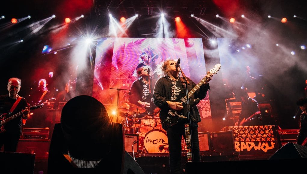 Robert Smith and The Cure performing their new album live in London. A guitarist with long grey-streaked hair and eyeliner sings and plays with his bandmates and a moody light show in the background.