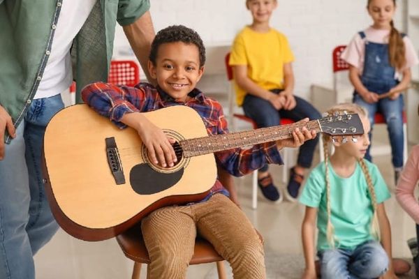 A little boy smiles and holds a guitar that is almost as big as he is.