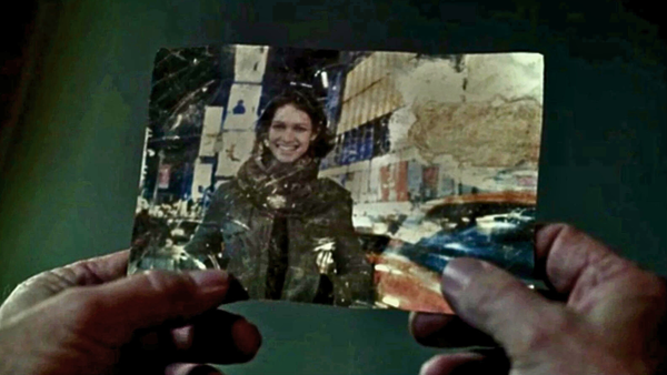 A pair of hands hold a crinkled photo of a smiling woman in a leather jacket against an urban backdrop.
