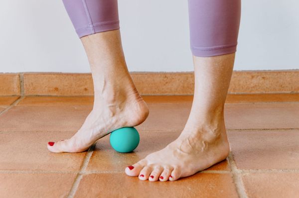 Two bare feet, the toenails painted bright red. Under the right foot is a small massage ball, demonstrating how to perform the plantar fascia release exercise described later in the column.