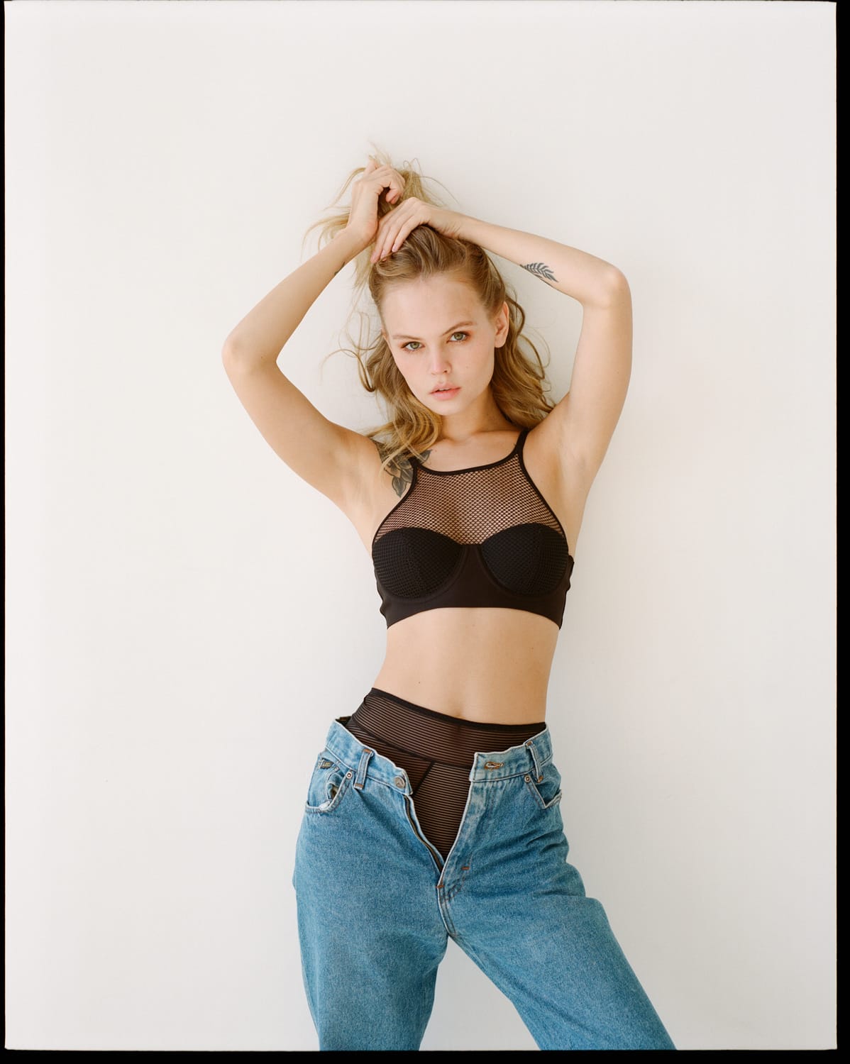Anastasiya Scheglova stands against a white wall with arms raised, wearing a black mesh halter top and blue jeans, photographed by Alina Korneeva.