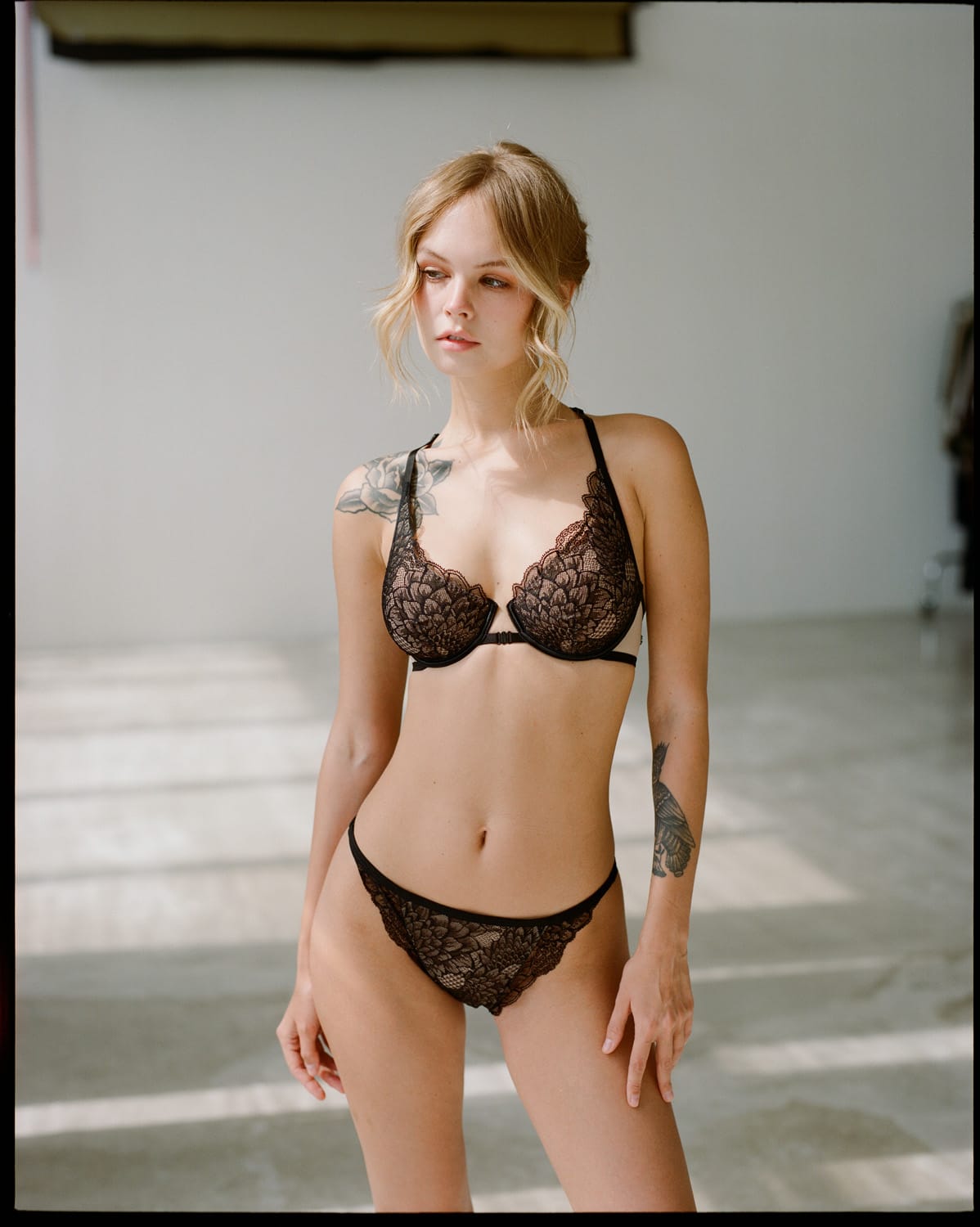 Anastasiya Scheglova stands in a black lace bra and briefs with window light behind her, photographed by Alina Korneeva.