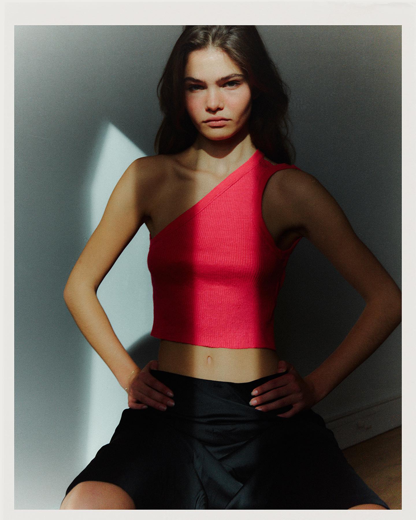 Anna faces the camera with hands on hips in a coral one-shoulder crop top and black skirt, lit by a window shaft, photographed by Ksenya Poggenpohl.