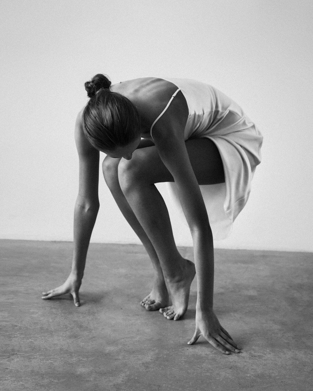 Olita Pimonova crouches low to the concrete floor in a white slip dress, face hidden, photographed by Maria Kudryavtseva.