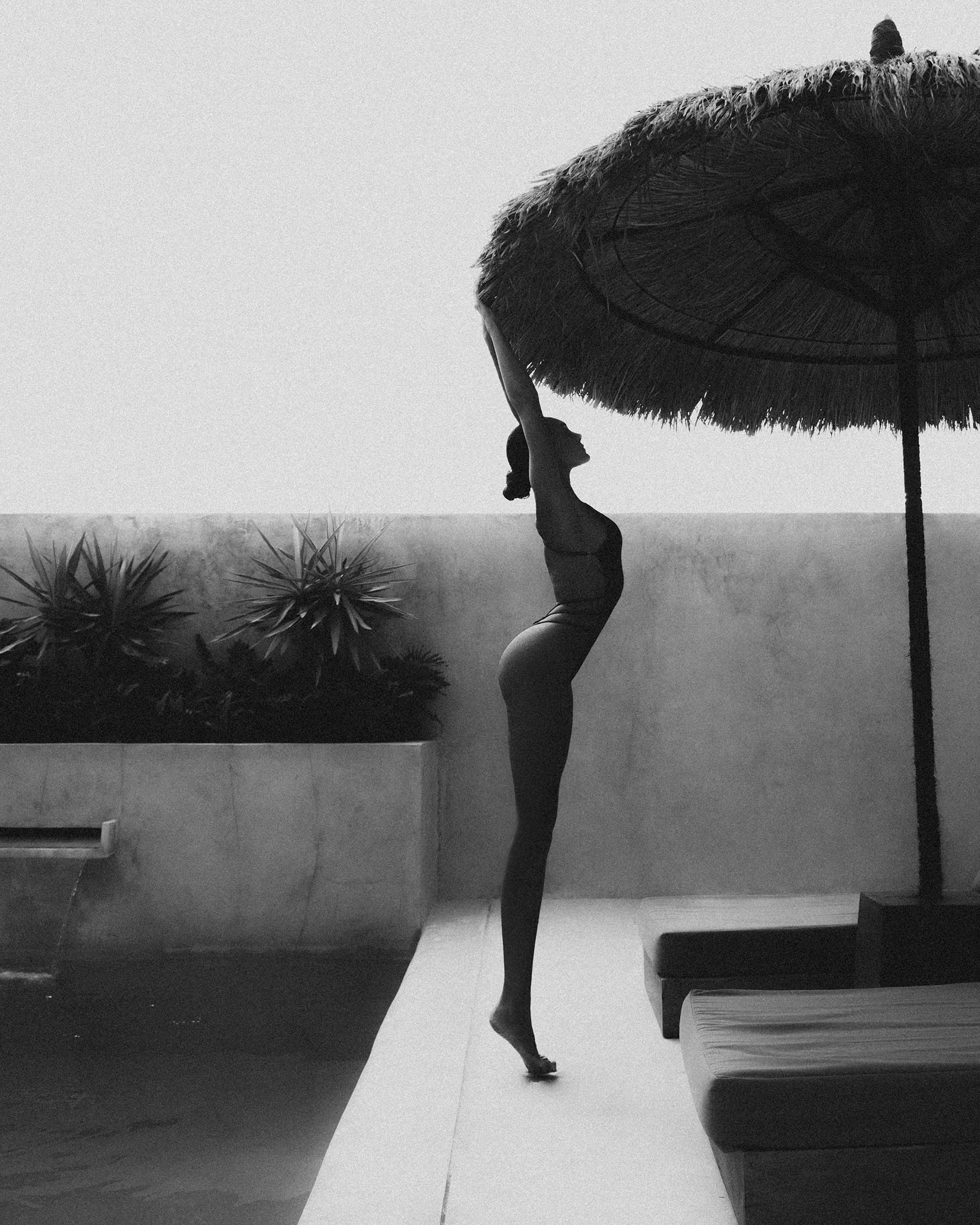 Sara Frazzetta reaches for a thatched palapa umbrella on tiptoes in a bikini, silhouetted against a pale sky, photographed by Tatyana Kudryashova.