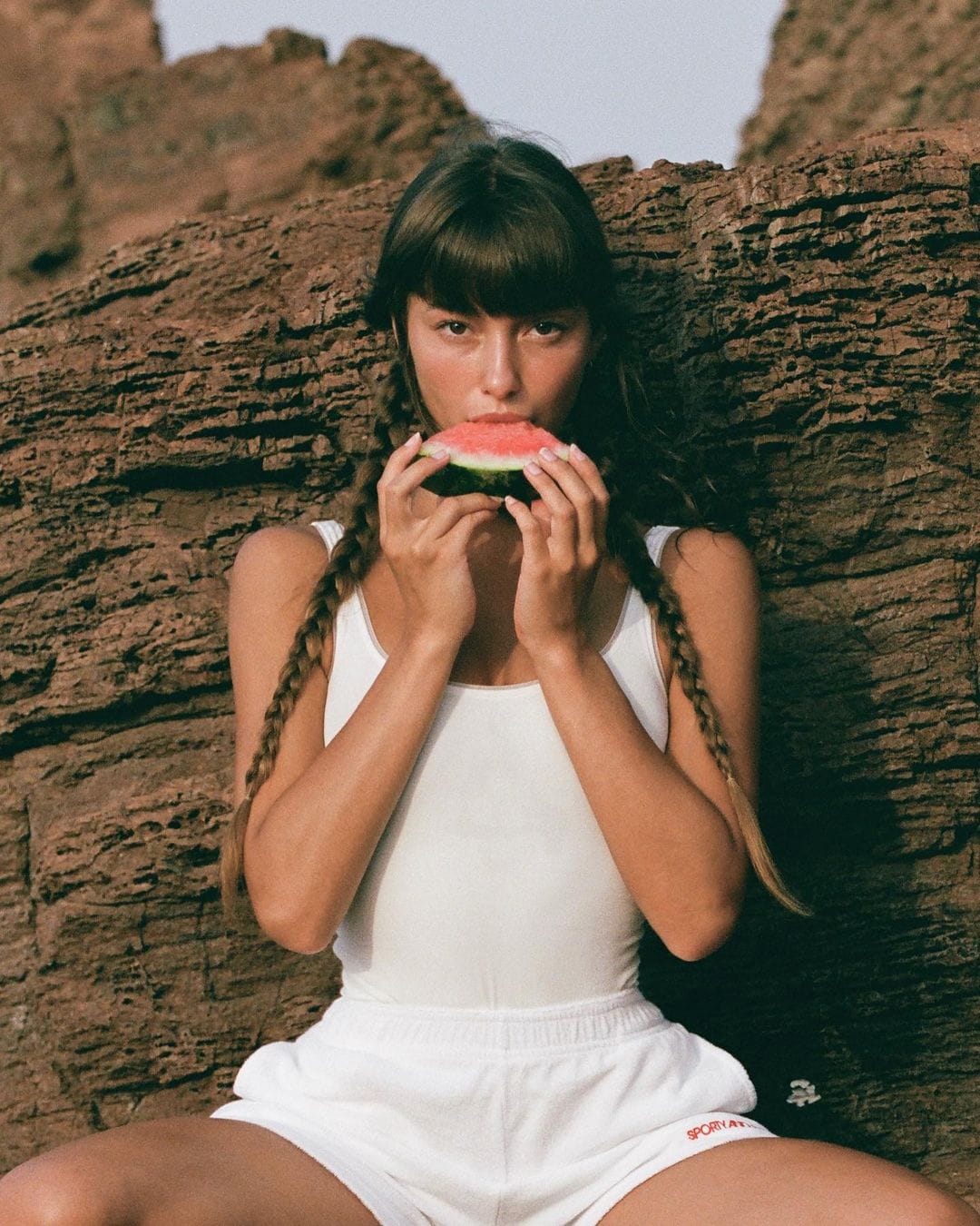 Telma Louise bites into a slice of watermelon against red-brown rock, wearing a one-piece and shorts with hair in braids, photographed by Anna Klembarova.