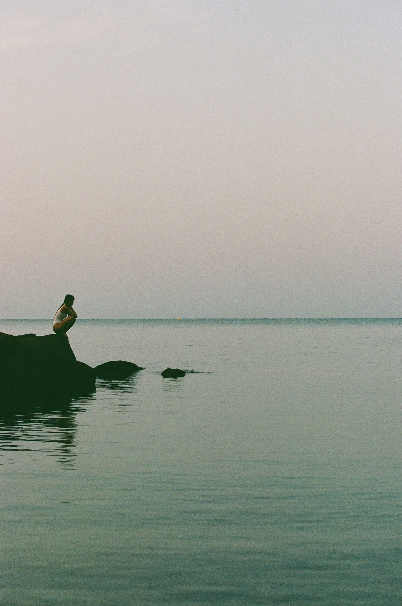 Telma Louise sits alone on a dark rock at the edge of a still sea, small in the frame against open water and sky, photographed by Anna Klembarova.