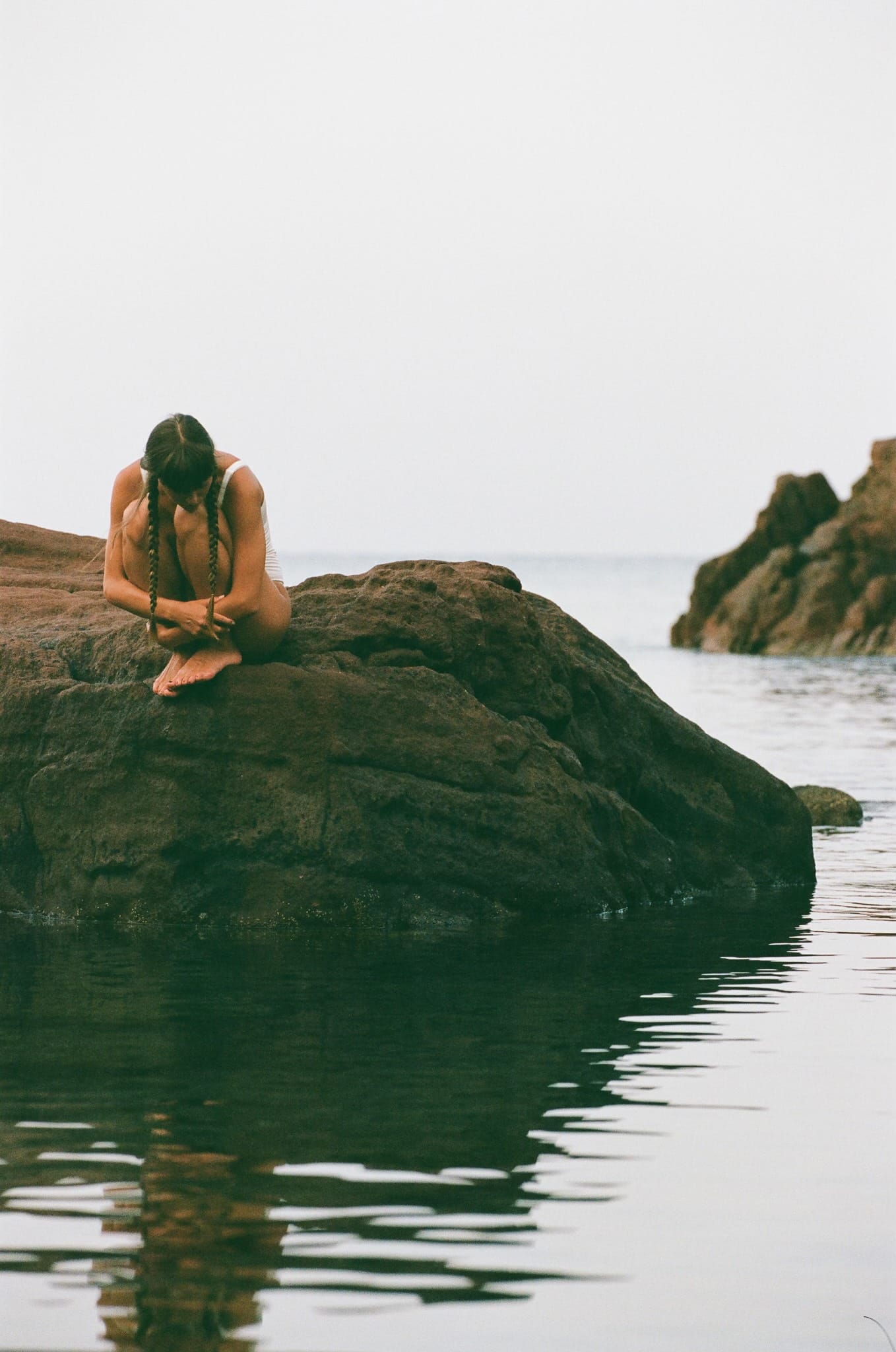 Telma Louise curls forward on a coastal rock with her head down, braids hanging, sea and rock formations behind her, photographed by Anna Klembarova.