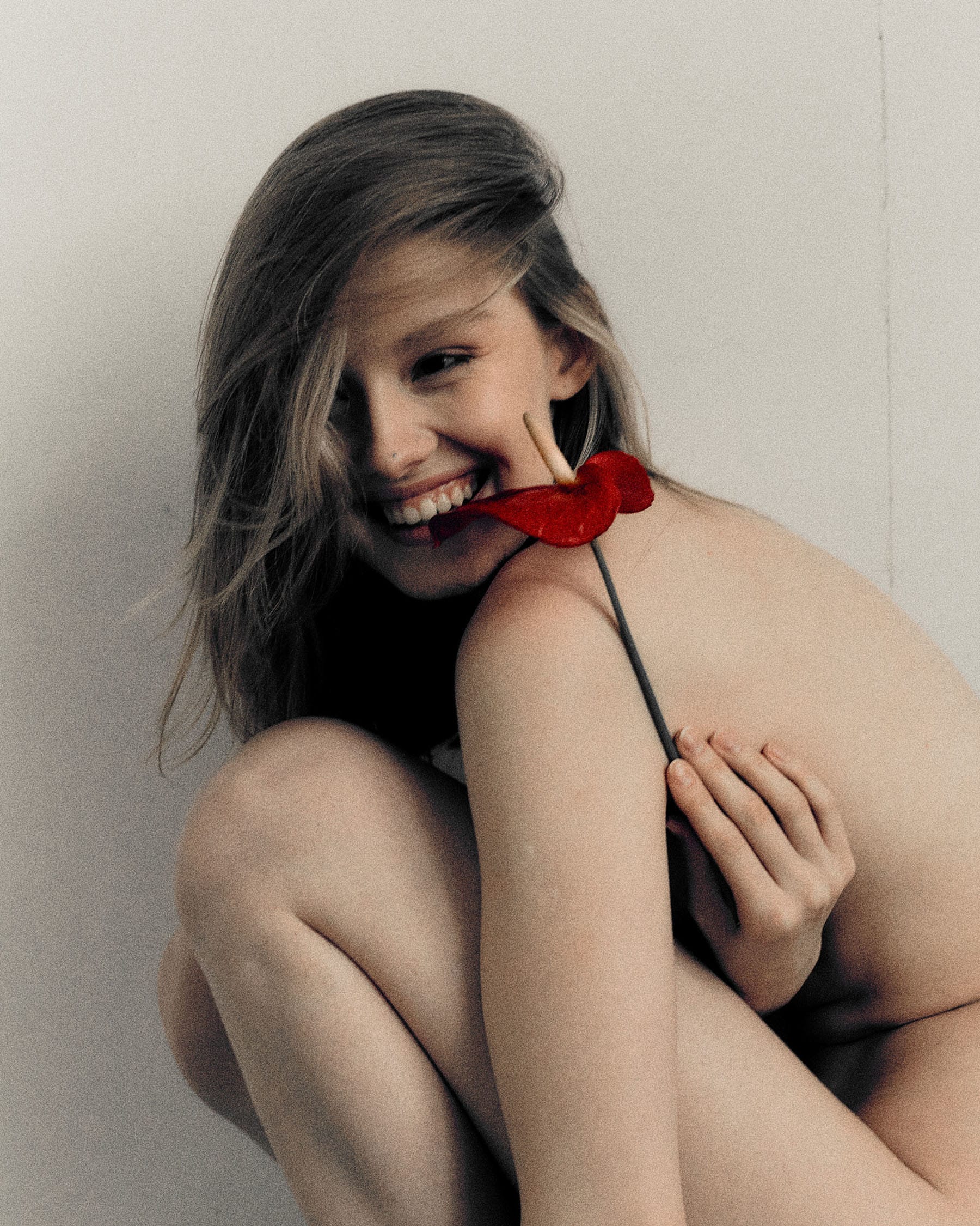 Camilla smiles with a red anthurium resting on her shoulder, knees drawn up, photographed by Ekaterina Kachevskaya.