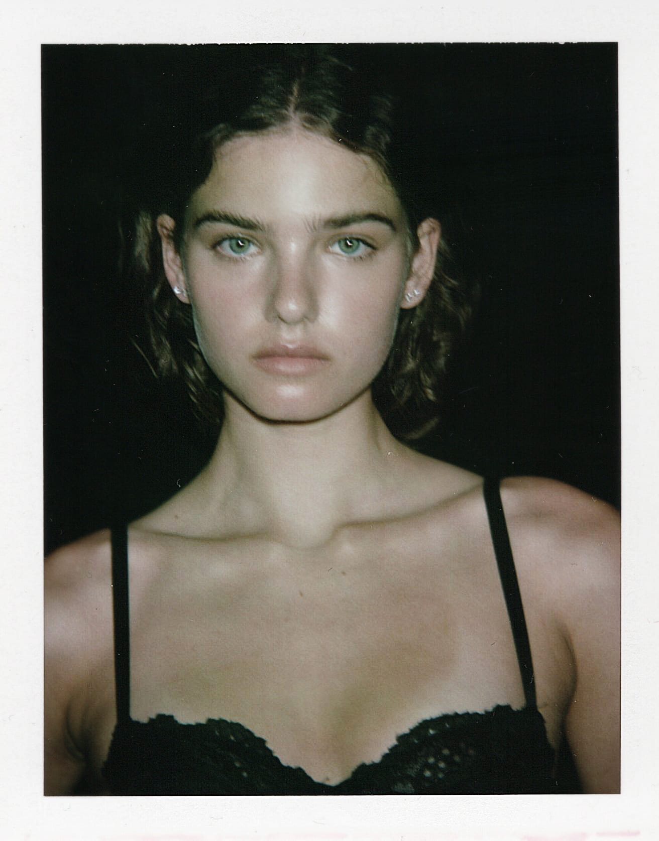 Close portrait of Maya Norman in a black lace camisole against a dark background, hair pulled back wet, shot on instant film by Bellezzamodaph.