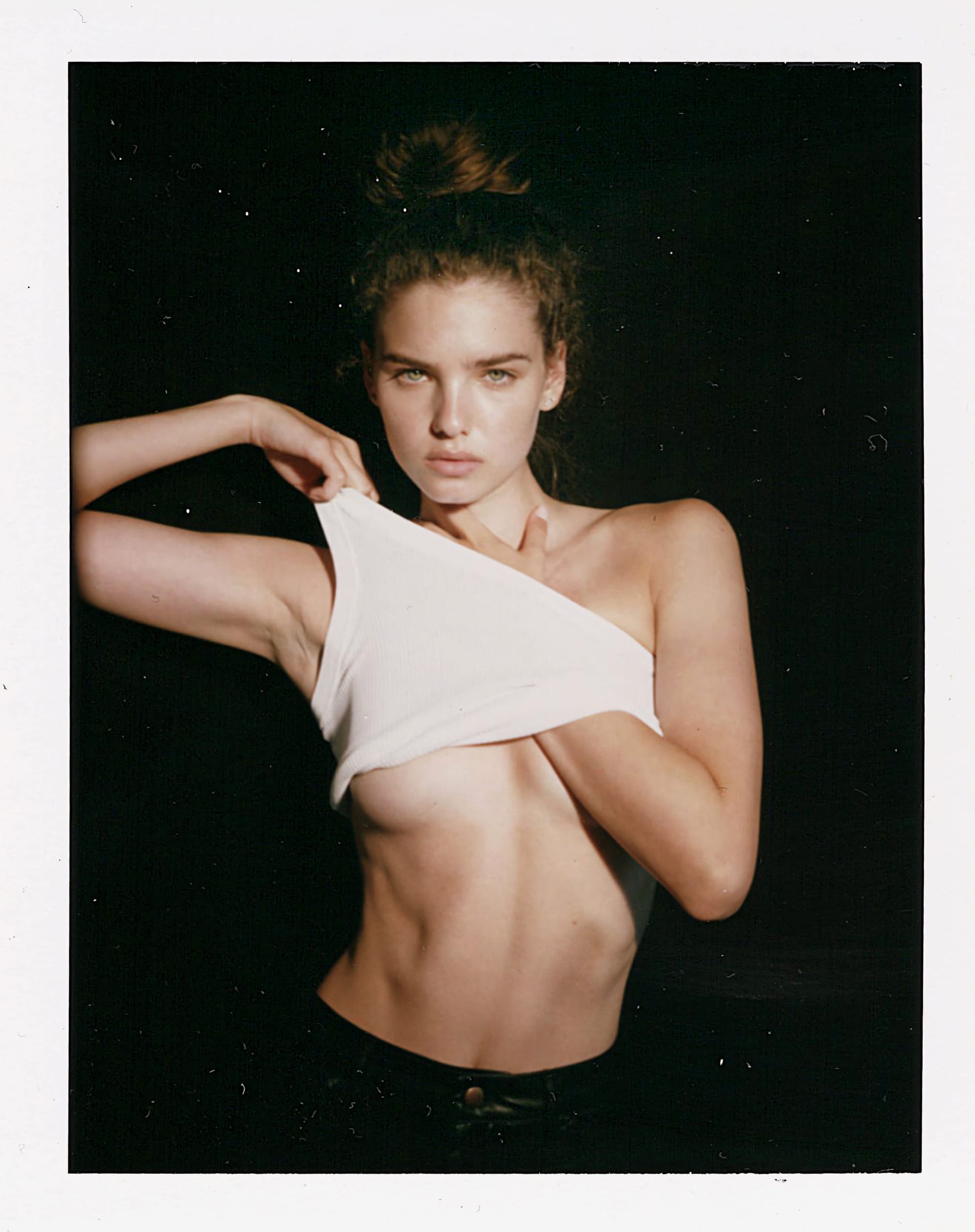 Maya Norman lifts a white ribbed tank to expose her underboob, hair in a messy bun, shot on instant film against black by Bellezzamodaph.