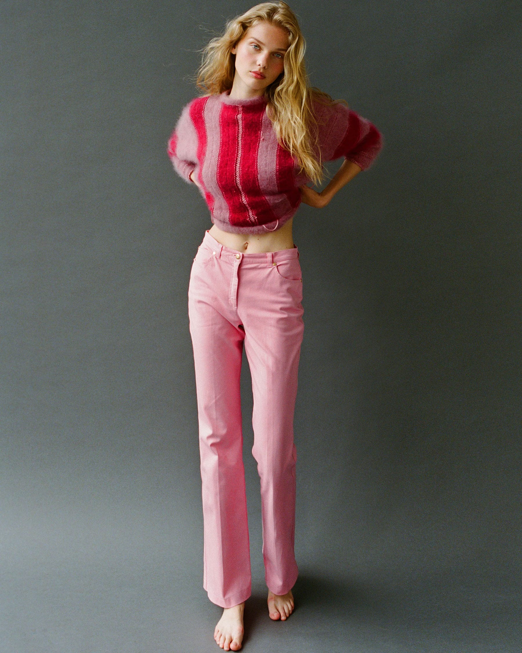 Vasilina Kireenko stands barefoot in a pink striped mohair sweater and pink trousers against a grey backdrop, photographed by Sasha Tsyganok.