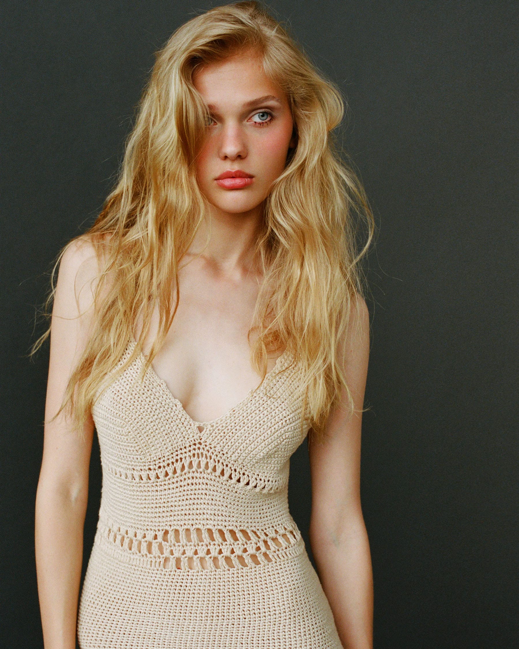 Vasilina Kireenko in a cream crochet dress with loose blonde hair, shot from the waist up against a dark background, photographed by Sasha Tsyganok.