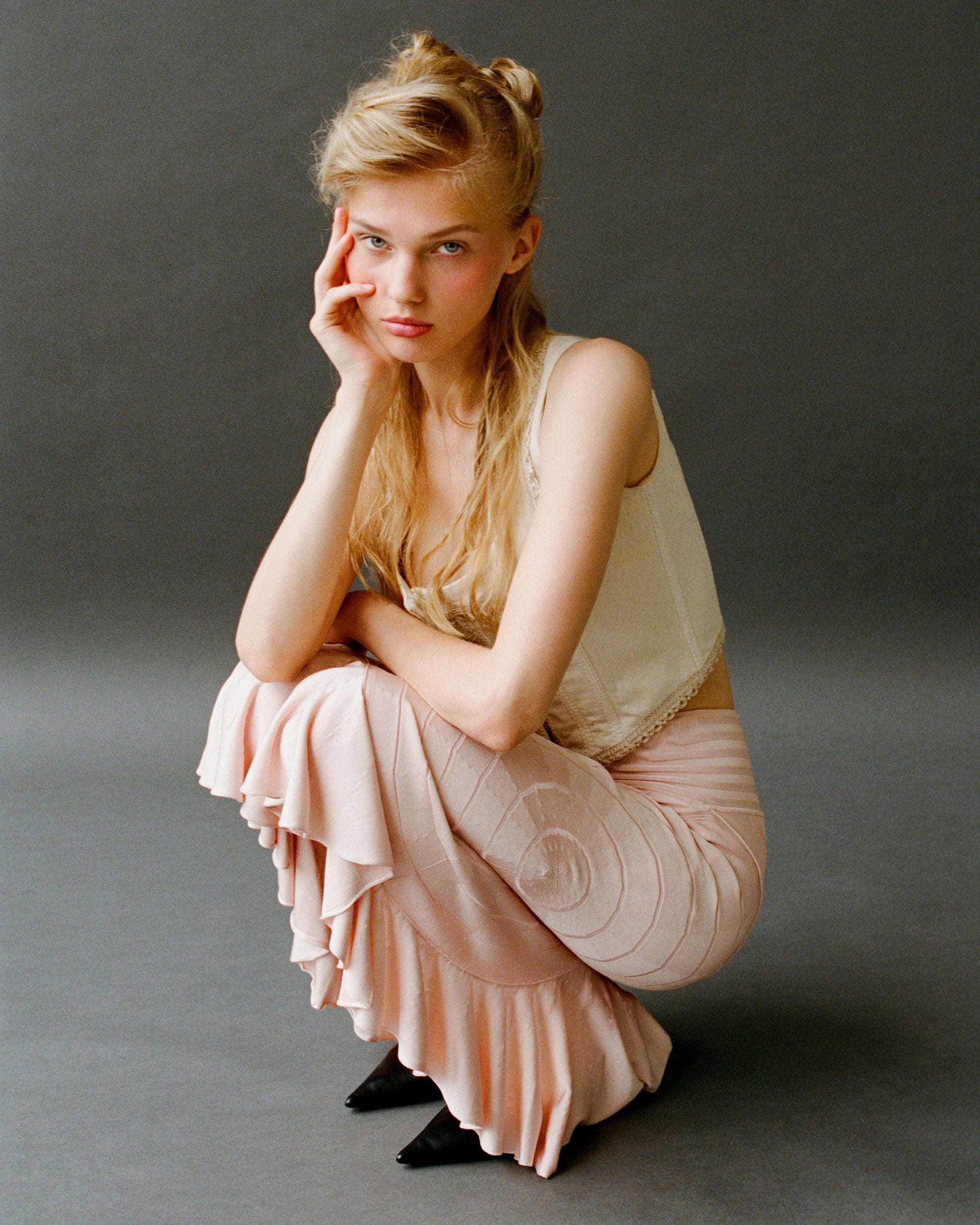 Vasilina Kireenko crouches in a cream silk top and pink ruffled skirt with black pointed shoes, hand to her face, photographed by Sasha Tsyganok.
