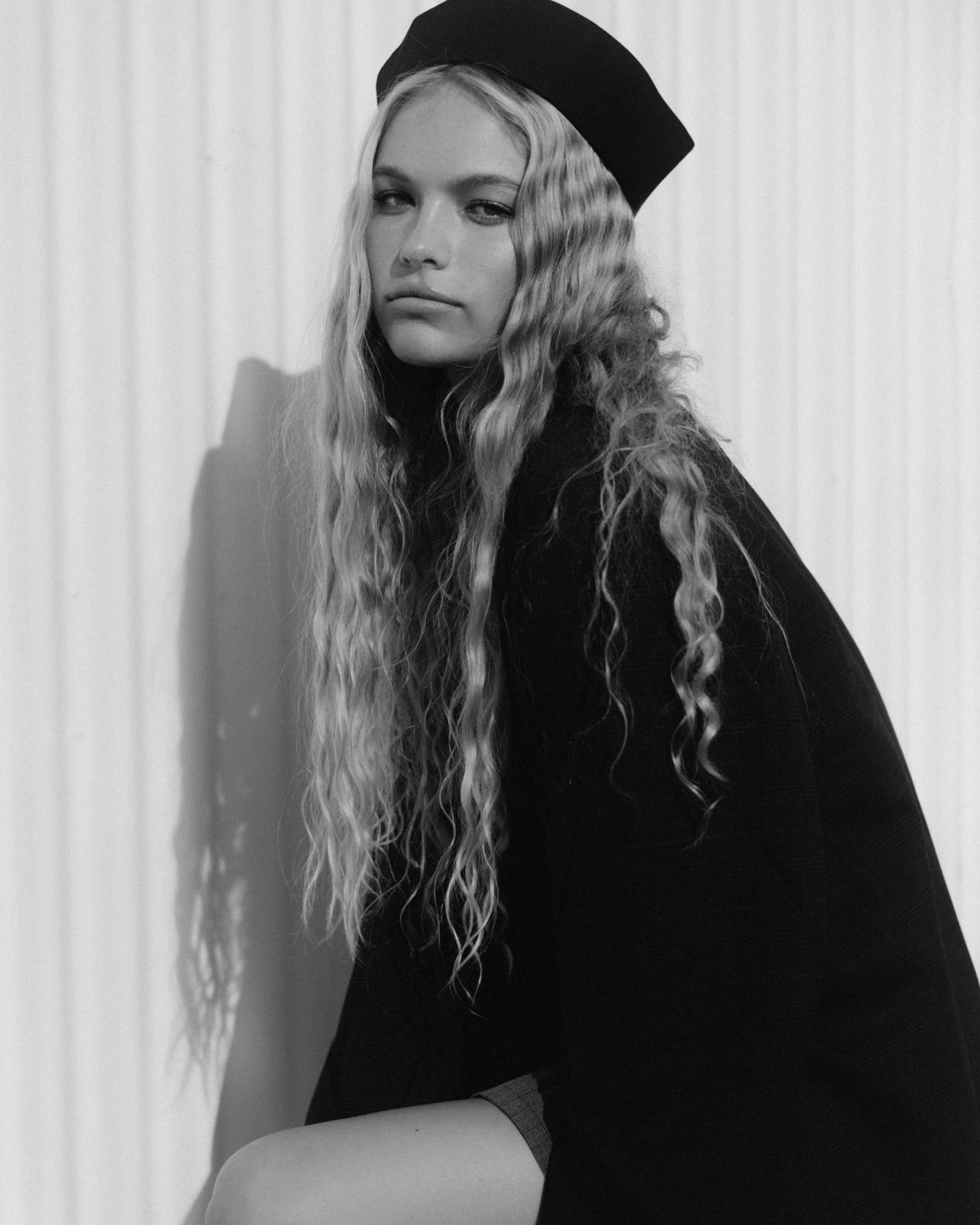 Cadence van Hoften in a black coat and pillbox hat against corrugated metal, long wavy hair over one shoulder, photographed in black and white by Olga Aleshina.