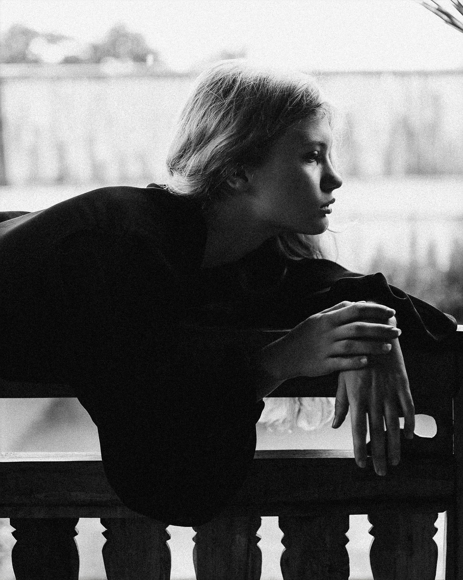 Eva leans over a veranda balustrade in a dark shirt, looking out past the camera, photographed in black and white by Tanya Xmyra.