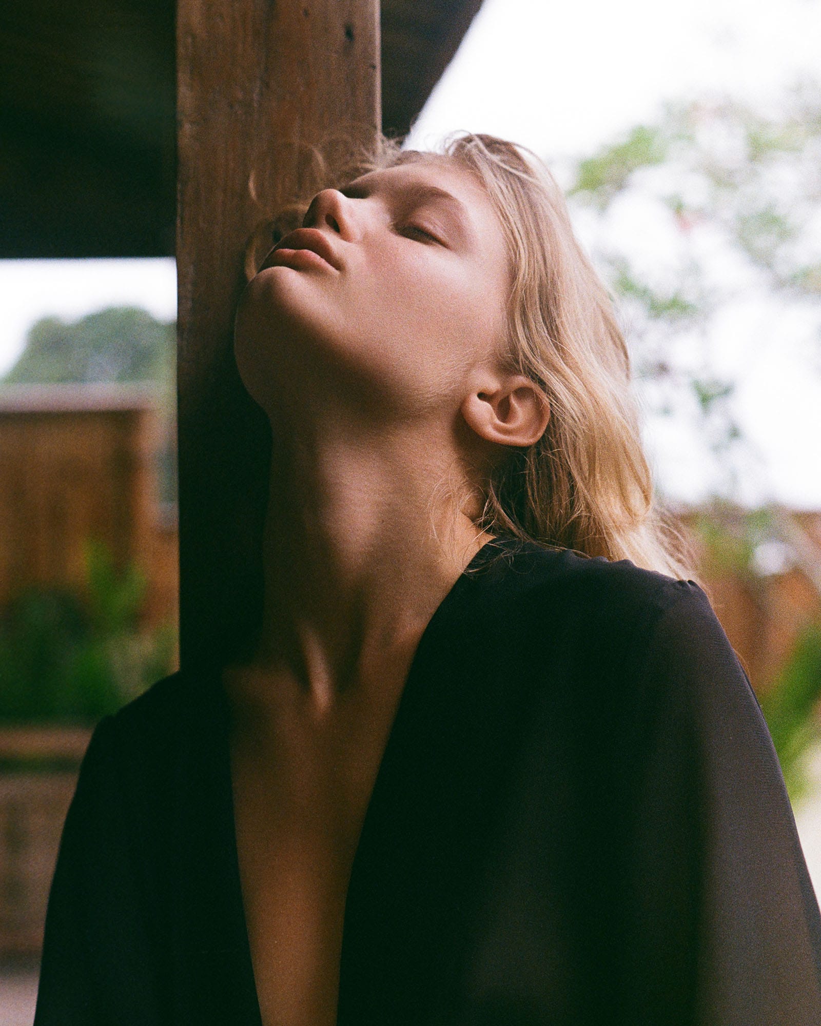 Close portrait of Eva leaning against a wooden post with head back and eyes closed in a dark open shirt, photographed by Tanya Xmyra.