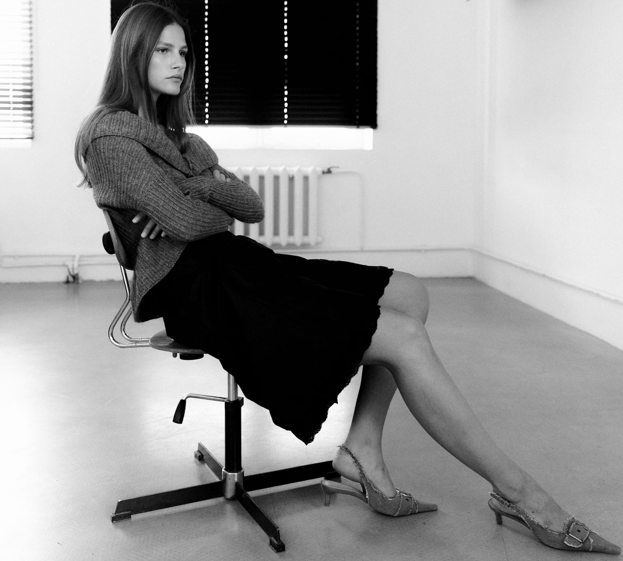 Eva Shevkoplyas slouches in a swivel chair in a ribbed sweater and black skirt with buckled heels, legs extended, photographed in black and white by Olesya Skvortsova.