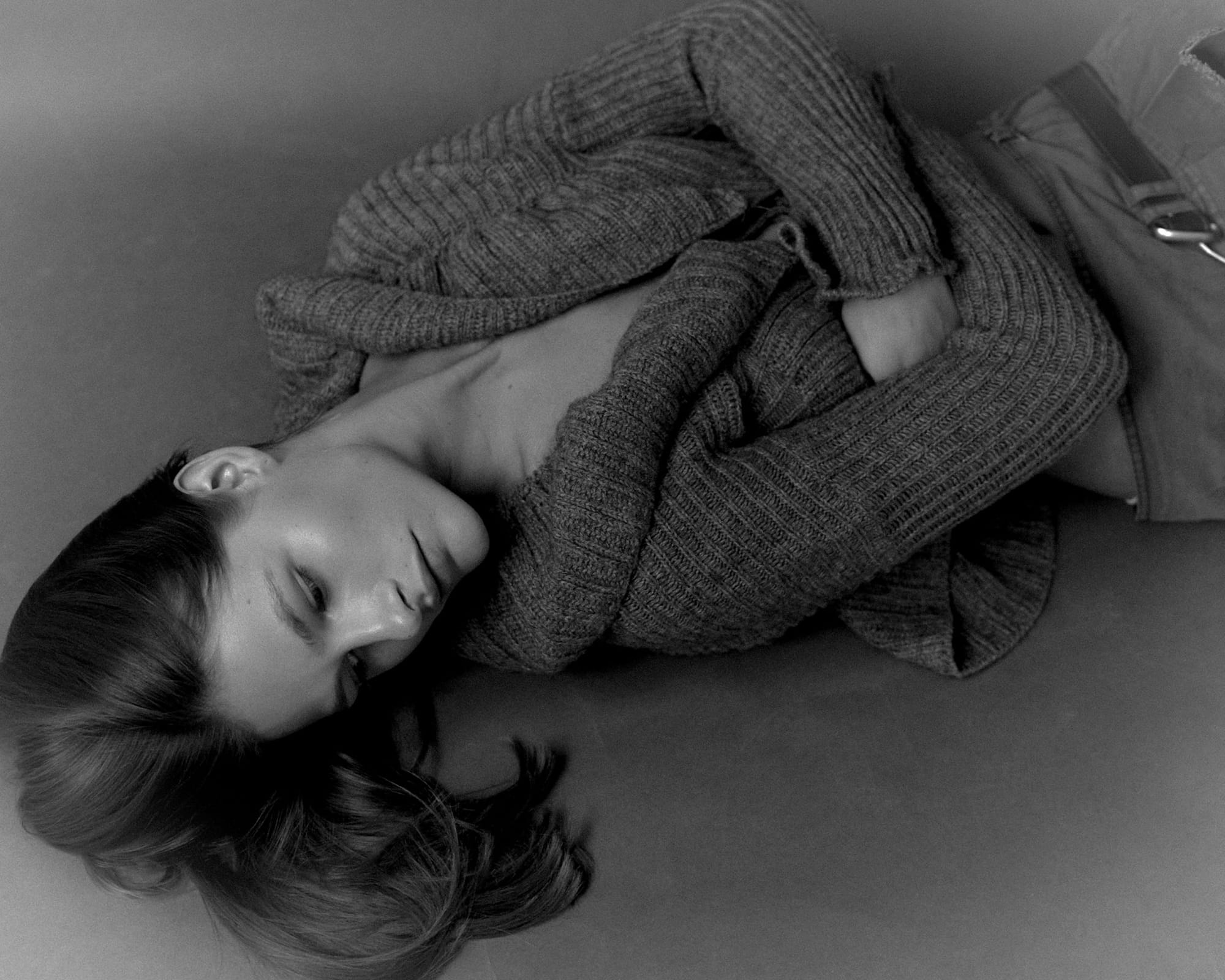 Eva Shevkoplyas lies on the studio floor in a ribbed cardigan, eyes downcast, hair fanning out, photographed in black and white by Olesya Skvortsova.