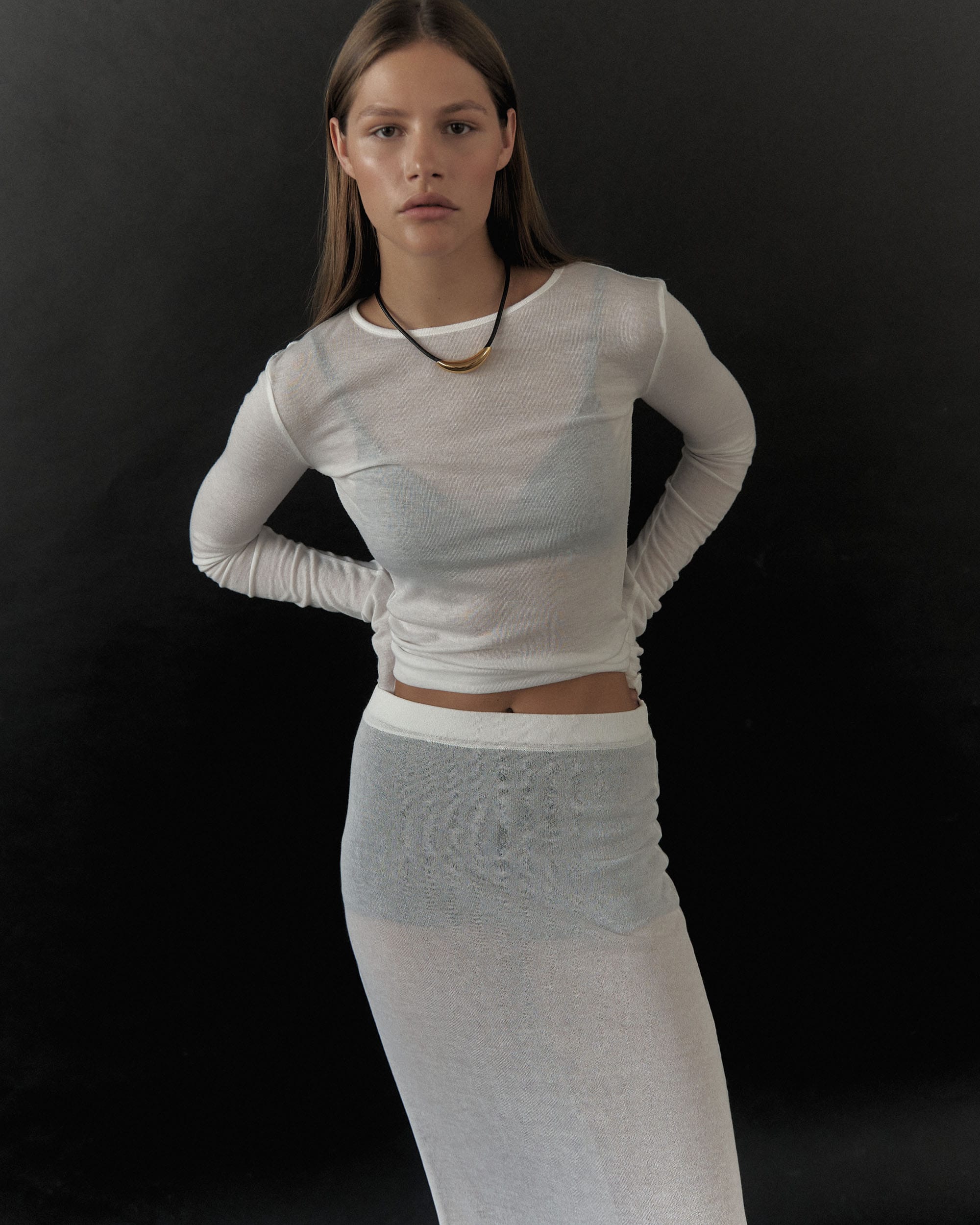 Eva Shevkoplyas stands in a sheer white top and matching skirt with a gold crescent necklace against a dark backdrop, photographed by Olesya Skvortsova.