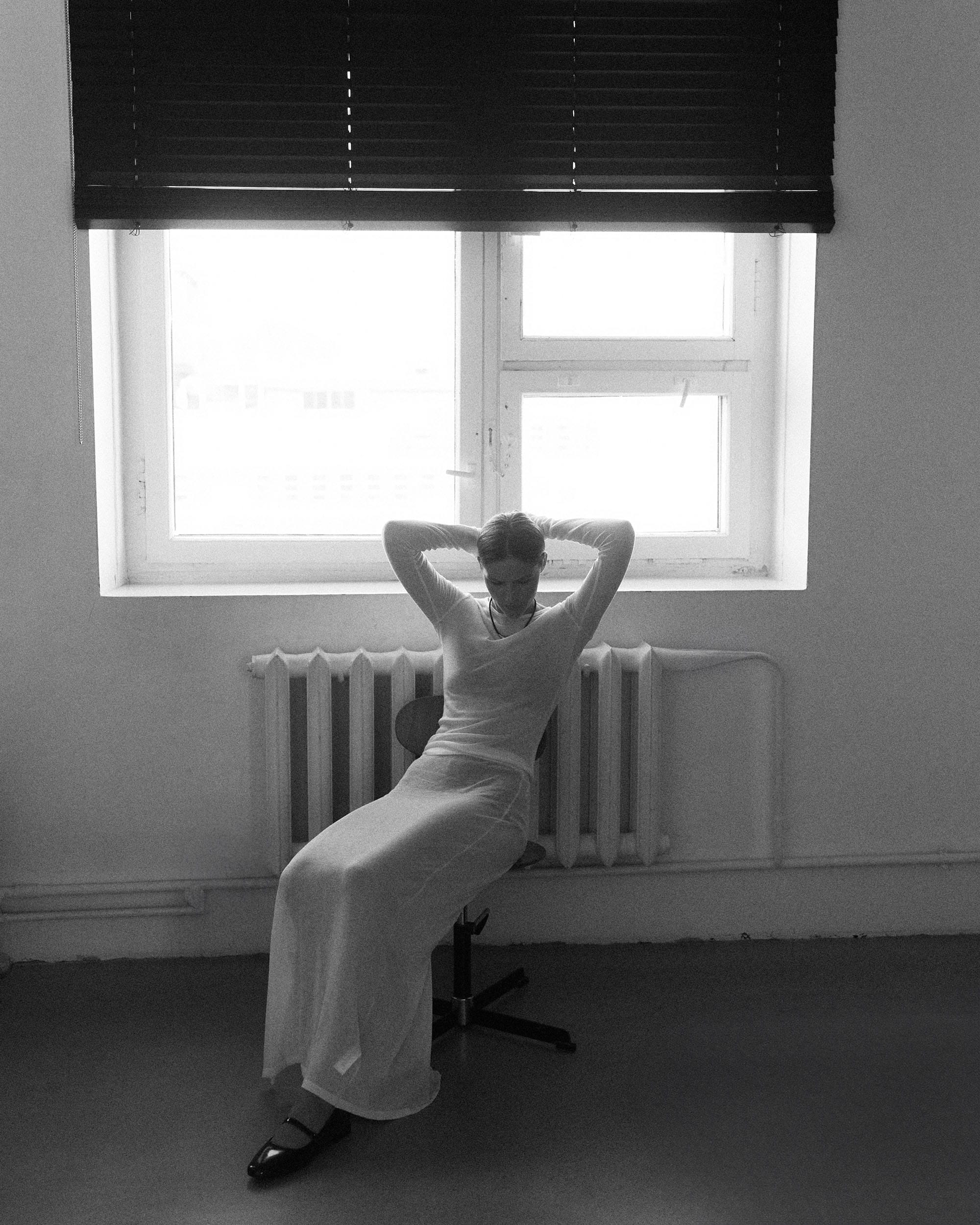 Eva Shevkoplyas sits on a swivel chair in a sheer white outfit, arms behind her head, radiator and blinds behind her, photographed by Olesya Skvortsova.