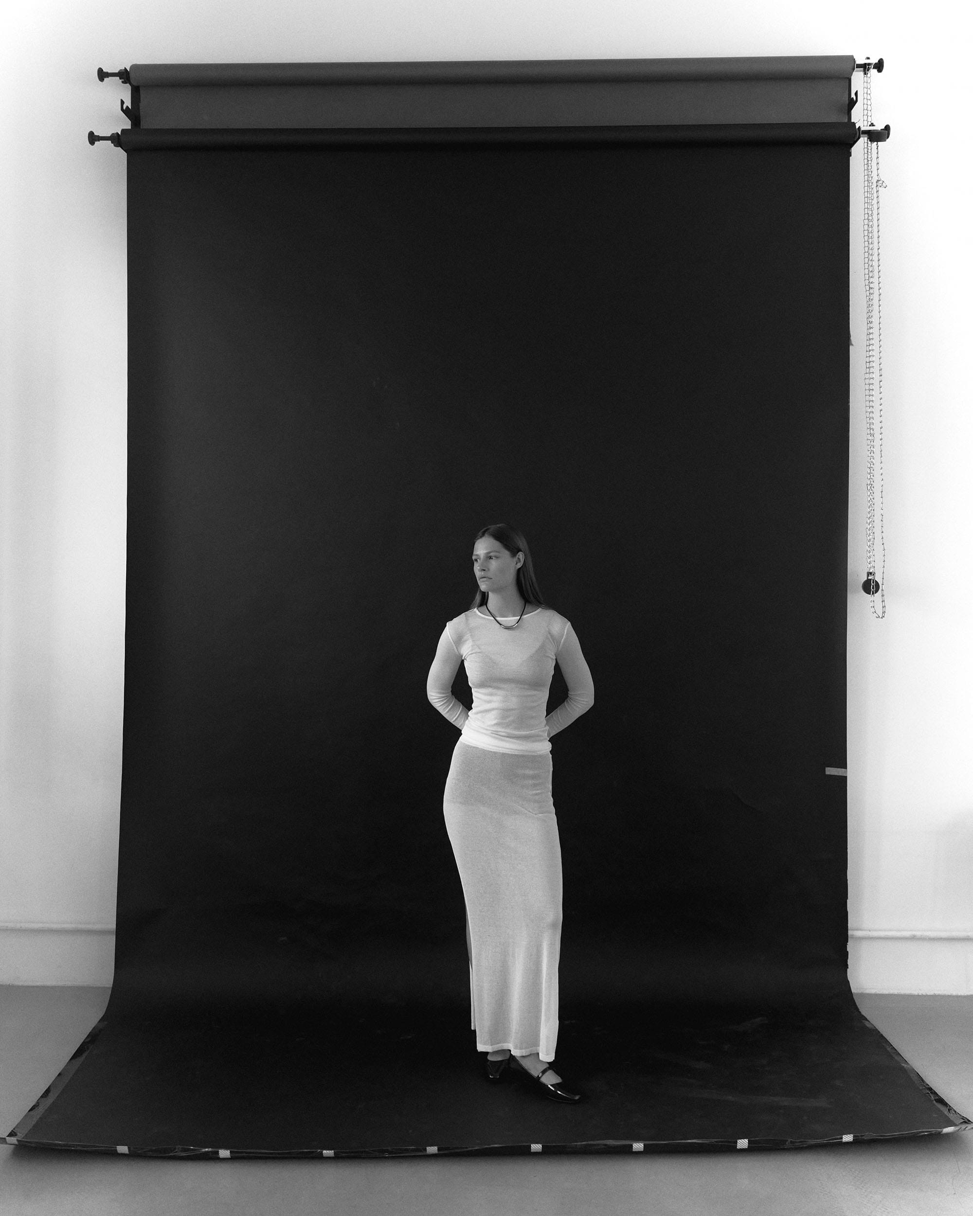 Eva Shevkoplyas stands centred against a pull-down black backdrop in a sheer white top and long skirt, photographed by Olesya Skvortsova.