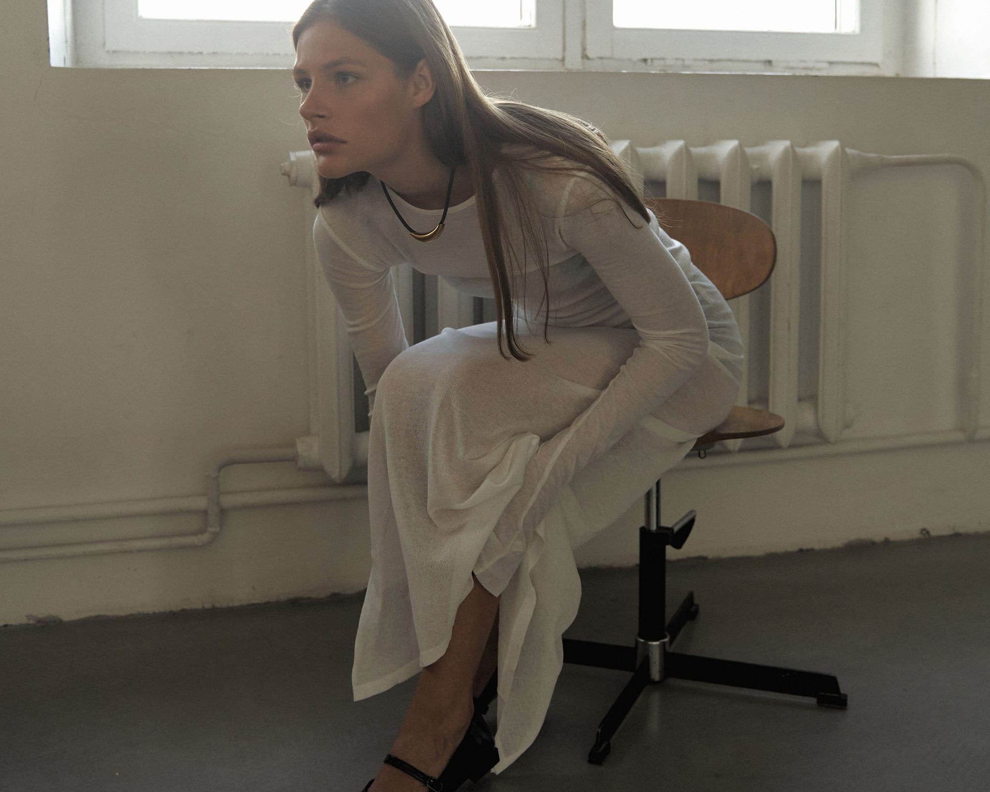 Eva Shevkoplyas leans forward on a swivel chair in a sheer white outfit and black heels, radiator behind her, photographed by Olesya Skvortsova.
