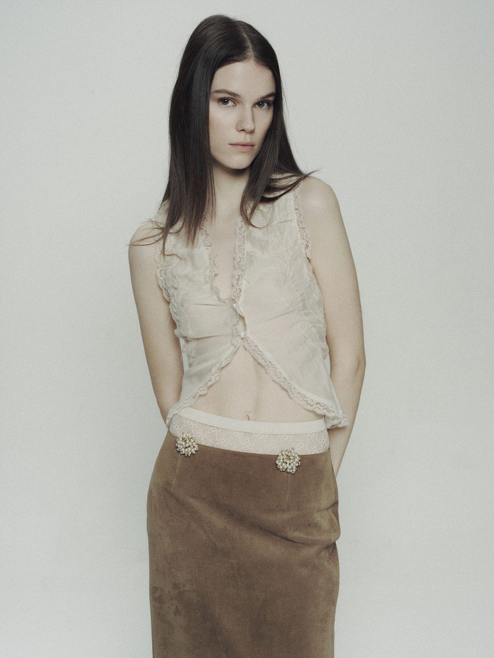 Greta Dethlefs in a cream lace top and brown suede skirt with pearl floret details, hands behind her back, photographed by Pascal Bräuer.