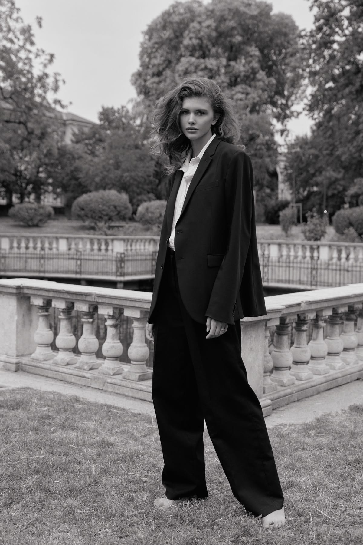 Alyona Lihosherstowa stands barefoot in grass in an oversized black suit and white shirt, with a marble balustrade and trees behind her, photographed by Alexei Bazdarev.