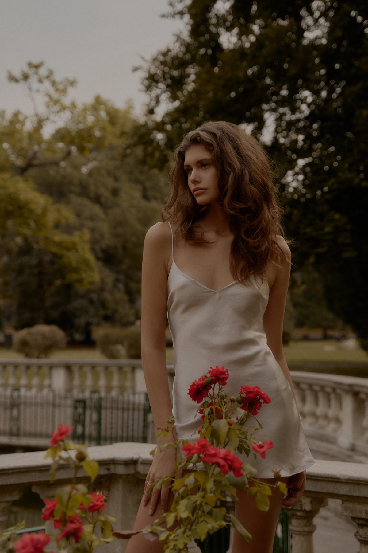 Alyona Lihosherstowa stands behind a cluster of red roses in an ivory slip dress, a park balustrade behind her, photographed by Alexei Bazdarev.