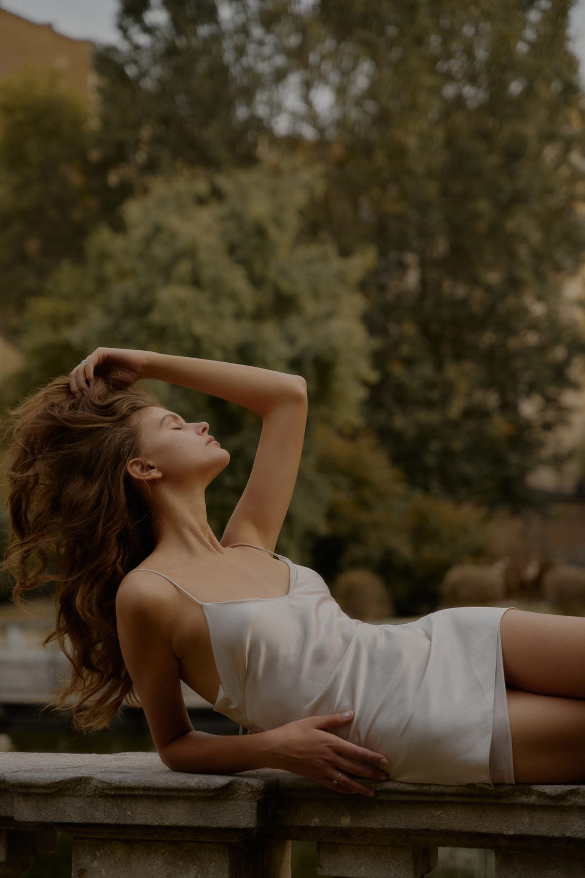 Alyona Lihosherstowa lies back on a stone surface with eyes closed and one hand in her hair, the ivory slip dress catching the light, photographed by Alexei Bazdarev.
