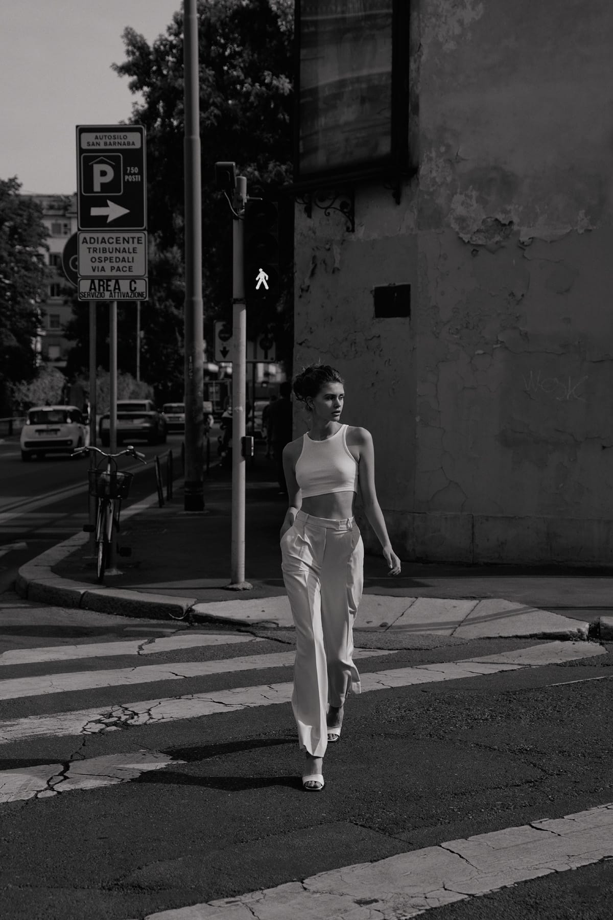 Alyona Lihosherstowa crosses a Milanese street near an Autosilo San Barnaba sign in a white crop top and cream wide-leg trousers, photographed by Alexei Bazdarev.